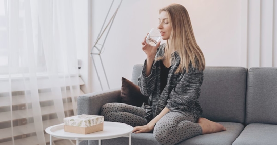 young woman with blonde hair sitting on the couch drinking a glass of water