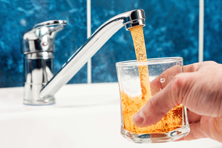 A hand holds a glass into which rusty dirty water is pouring from a tap